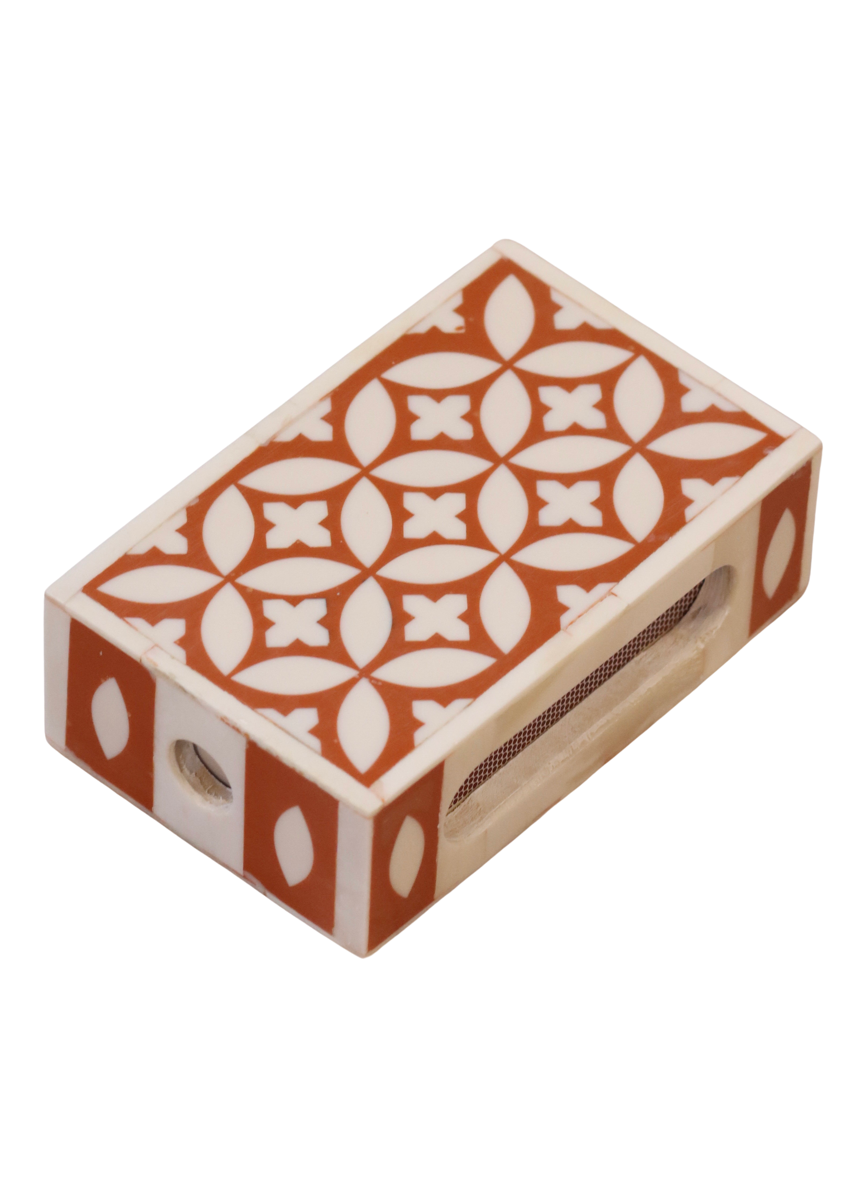 A handmade matchbox holder with a geometric pattern in orange and white colors, equipped with a slot to hold matches.